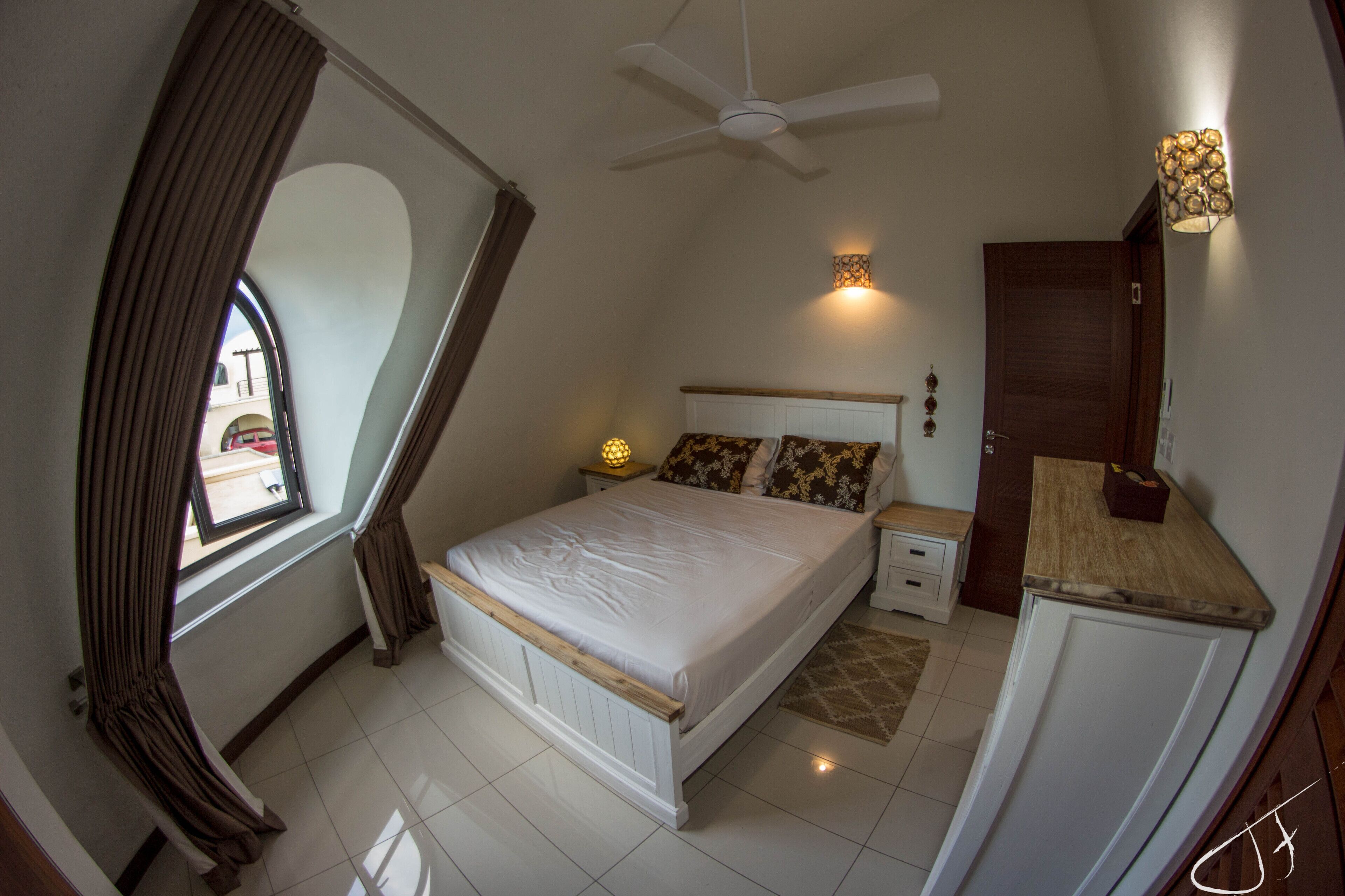 A bedroom with a bed, nightstands, a ceiling fan, a window with curtains, a wall-mounted light, and a door.