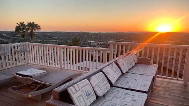 A wooden deck with a sofa and lounge chairs, overlooking a cityscape at sunset. - Beach Vibes (Spring Valley)