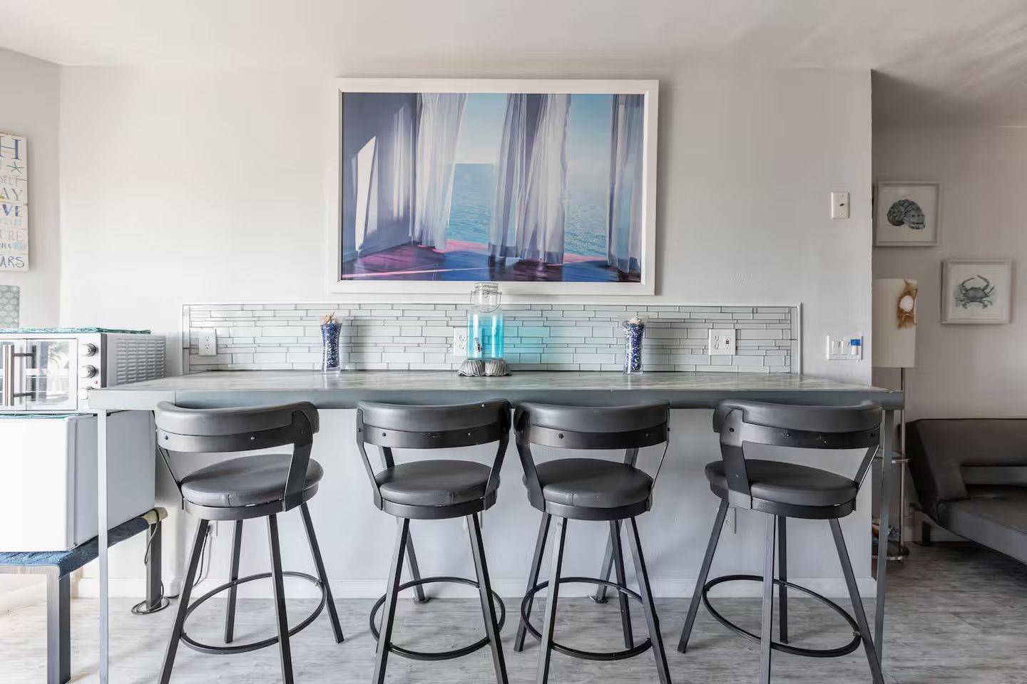 A modern kitchen with a bar area, featuring a large window with a view of the sea, a white tiled backsplash, and a framed artwork above the counter.
