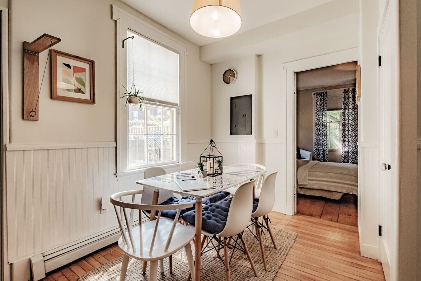 A dining area with a table, chairs, a hanging plant, a clock, and a view into another room.