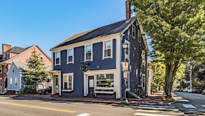 A two-story building with a dark blue exterior, a front entrance with a bench, and a street view with a crosswalk and parked cars. - Spacious West End Retreat, 3BR/2BA, Walk Everywhere + Private Deck (Portland)