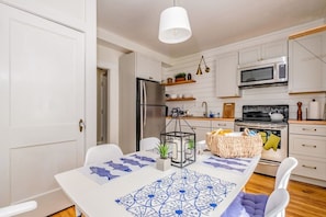 Cozinha moderna com armários brancos, eletrodomésticos de aço inoxidável e uma área de refeições com toalha de mesa estampada.