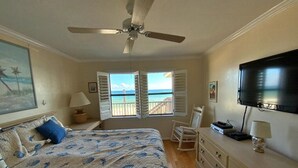 Un dormitorio con cama, ventilador de techo, cómoda, televisor y vista al océano a través de contraventanas abiertas.
