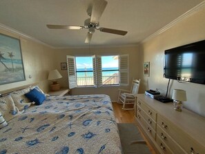 Ein Schlafzimmer mit Bett, Deckenventilator, Schminktisch, Fernseher und Meerblick durch geöffnete Fensterläden.
