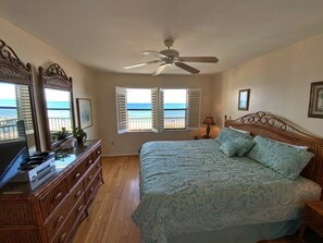 A bedroom with a bed, a dresser, a ceiling fan, and a view of the ocean through open windows.