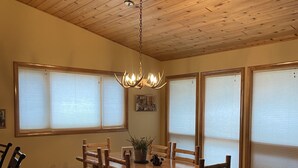 A dining area with a wooden table and chairs, a chandelier, and large windows with blinds.