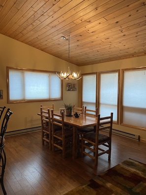 Un comedor con mesa y sillas de madera, una araña y ventanas grandes con persianas.