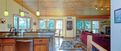 Cocina amplia con armarios de madera, encimera de granito y una isla grande. Hay un comedor con sofá rojo y chimenea.