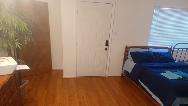A bedroom with a wooden floor, a white door, a bed with blue bedding, and a plant.