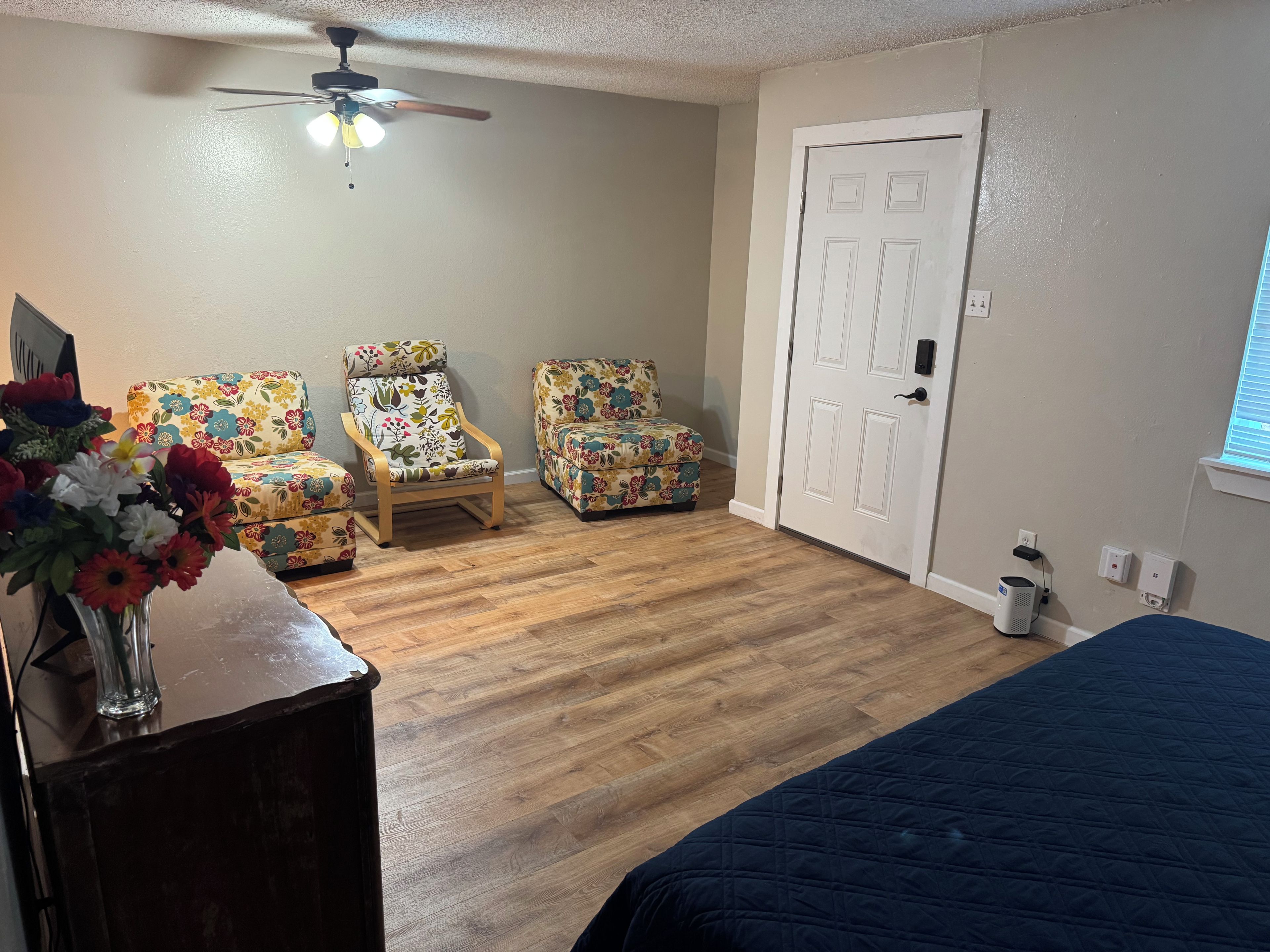 A bedroom with a bed, two floral armchairs, a ceiling fan, and a white door.