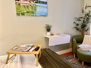 A room with a bed, a small wooden table with magazines and remotes, a green armchair, a white shelf with a plant, and a painting on the wall.