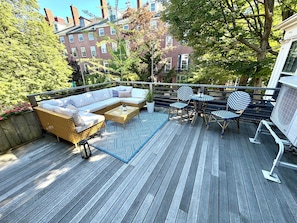 Una terraza con sofá, sillas y una mesa, rodeada de vegetación y edificios al fondo.