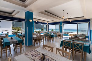 Un restaurante con mesas y sillas, amplias ventanas con vista al mar, y una decoración azul y blanca.