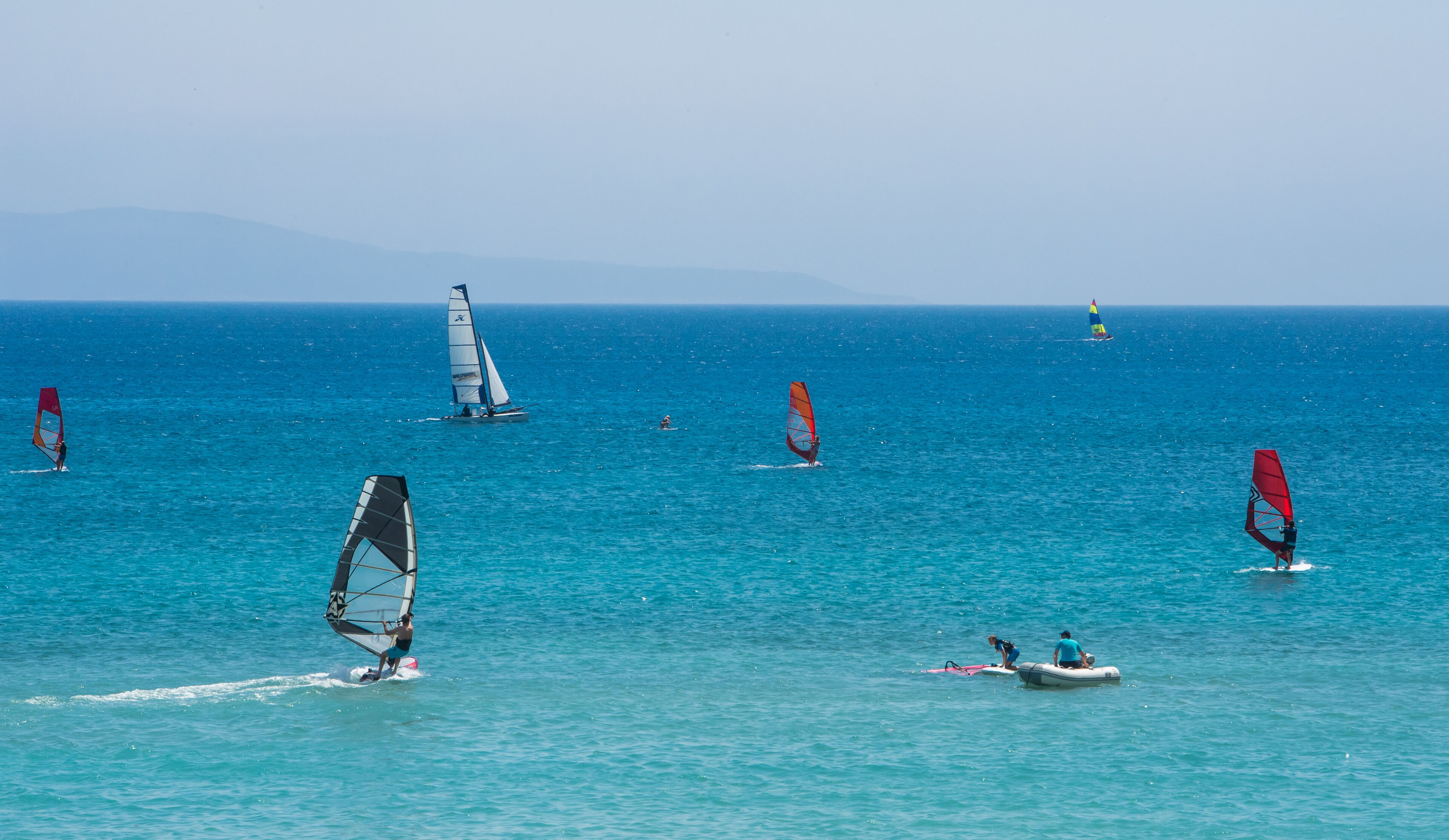 A coastal area with multiple windsurfers and a sailboat on the water.