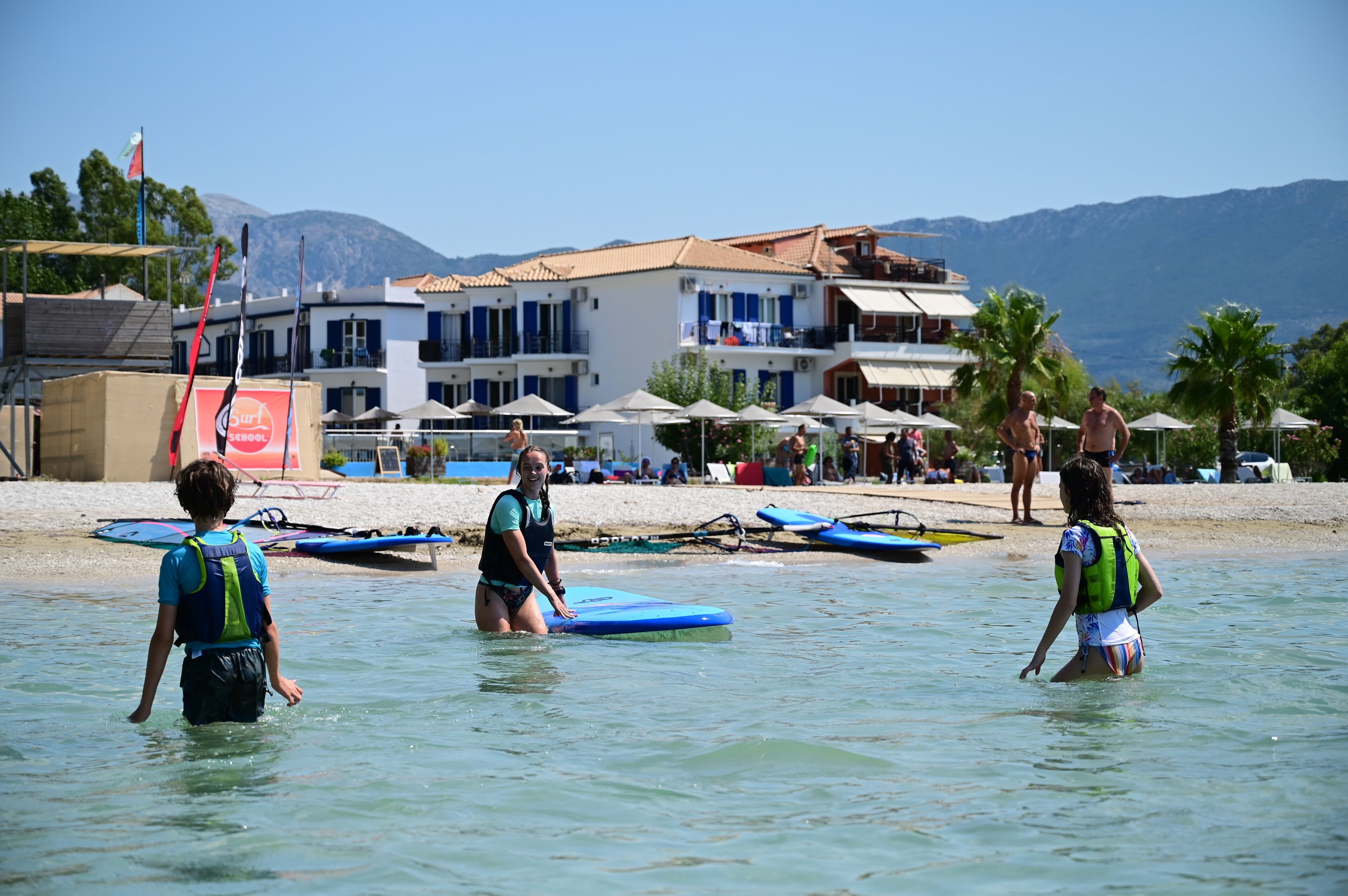 Un hotel frente a la playa con varios balcones, una playa de arena y una vista despejada de las montañas al fondo.