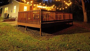 Une terrasse en bois illuminée de guirlandes lumineuses, une maison en arrière-plan et des arbres tout autour.
