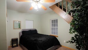 Une chambre à coucher comprenant un lit, un ventilateur de plafond, une fenêtre avec des stores, une petite table de chevet et une plante en pot.