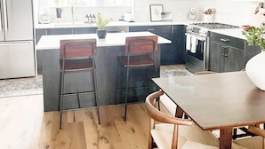 A modern kitchen with white cabinets, a central island with bar stools, a dining table, and a large window providing natural light.