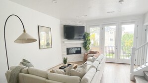 A modern living room with a beige sofa, a floor lamp, a flat-screen TV, and a staircase leading to an outdoor area.