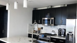 Une cuisine moderne avec des meubles de couleur sombre, un plan de travail en marbre, des appareils électroménagers en acier inoxydable et des luminaires suspendus.