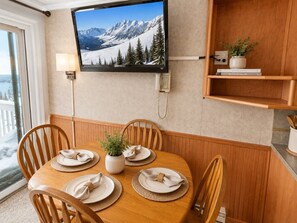 Un área de comedor con una mesa de madera preparada para cuatro personas, un televisor montado y un teléfono en la pared.
