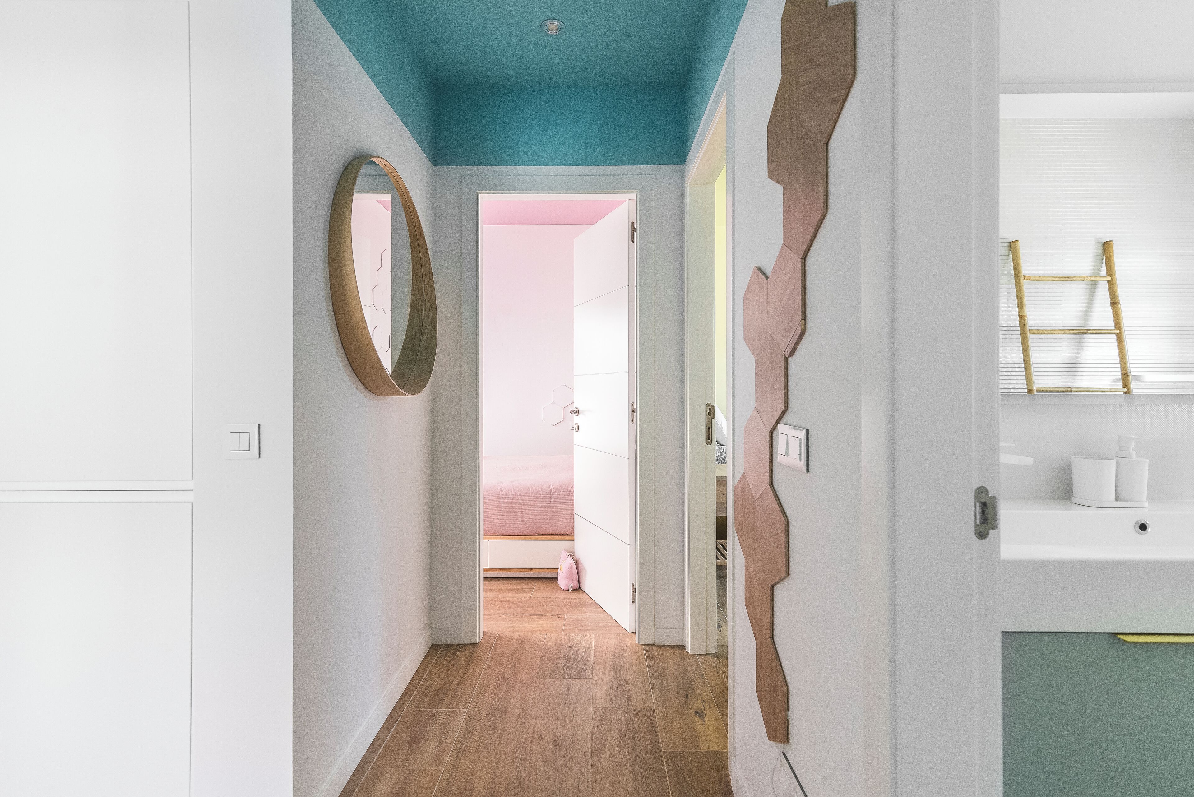 Un couloir doté d’un plancher en bois, d’un plafond de couleur turquoise et d’une porte donnant sur une pièce où se trouve un lit rose.