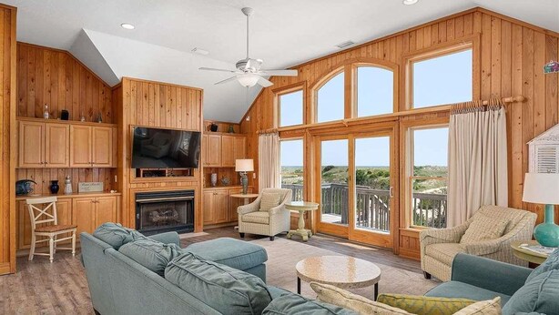 A spacious living room with a fireplace, wooden walls, a large TV, and a balcony with a view. - Oceanfront, Pet- & Handicap- Friendly Home w/ Heated Pool, Elevator & Game Room (Salvo)
