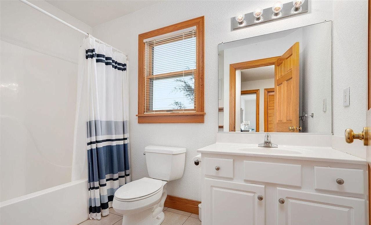 A bathroom with a white toilet, a bathtub with a blue and white striped shower curtain, a window with blinds, and a vanity with a mirror.