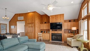 A spacious living room with a large sofa, a dining area with a table and chairs, wooden paneling, a fireplace, a television, and a ceiling fan. - Oceanfront, Pet- & Handicap- Friendly Home w/ Heated Pool, Elevator & Game Room (Salvo)