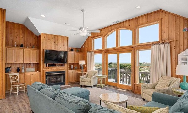 A spacious living room with a fireplace, wooden walls, a large TV, and a balcony with a view. - Oceanfront, Pet- & Handicap- Friendly Home w/ Heated Pool, Elevator & Game Room (Salvo)