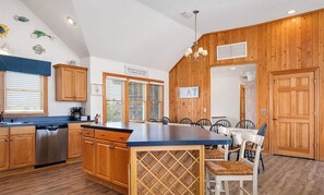 A kitchen with wooden cabinets, a central island with a wine rack, and a dining area with chairs. - Oceanfront, Pet- & Handicap- Friendly Home w/ Heated Pool, Elevator & Game Room (Salvo)