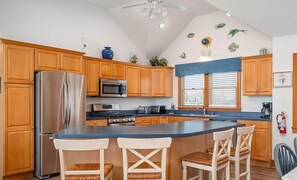 A kitchen with wooden cabinets, stainless steel appliances, a central island with bar stools, and decorative fish wall art. - Oceanfront, Pet- & Handicap- Friendly Home w/ Heated Pool, Elevator & Game Room (Salvo)