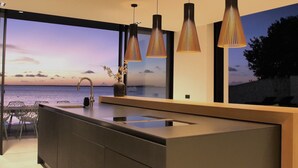 Une cuisine moderne dotée d’un grand îlot central, de luminaires suspendus et offrant une vue sur la mer.