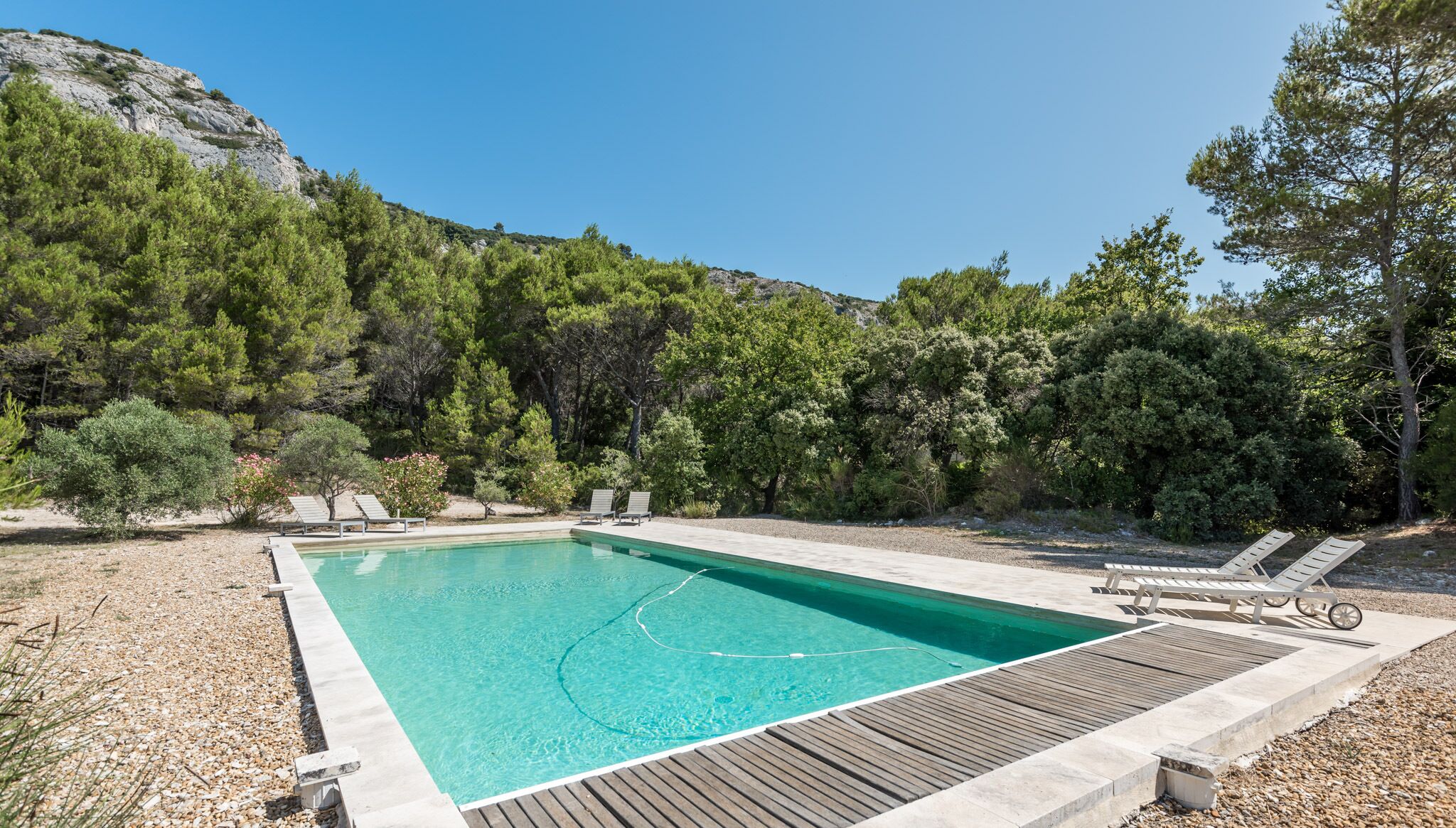 A rectangular pool with a wooden deck and lounge chairs, surrounded by trees and a rocky hillside.