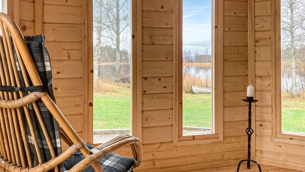 A wooden interior with a wicker chair, a candle holder, and a view of a lake through large windows. - Stunning home in Svanskog with sauna (Svanskog)