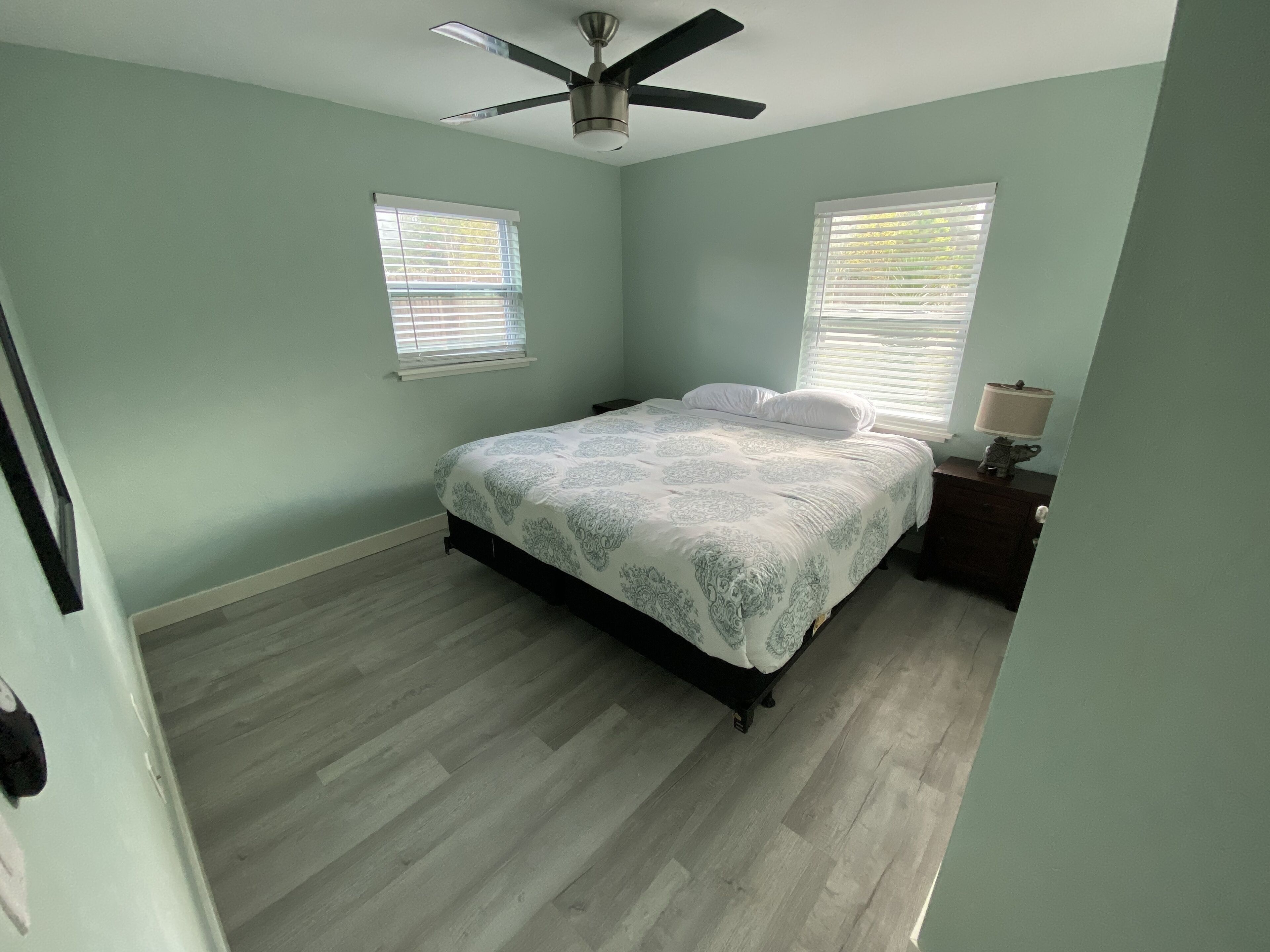 A bedroom with a bed, two windows with blinds, a ceiling fan, and a nightstand with a lamp.