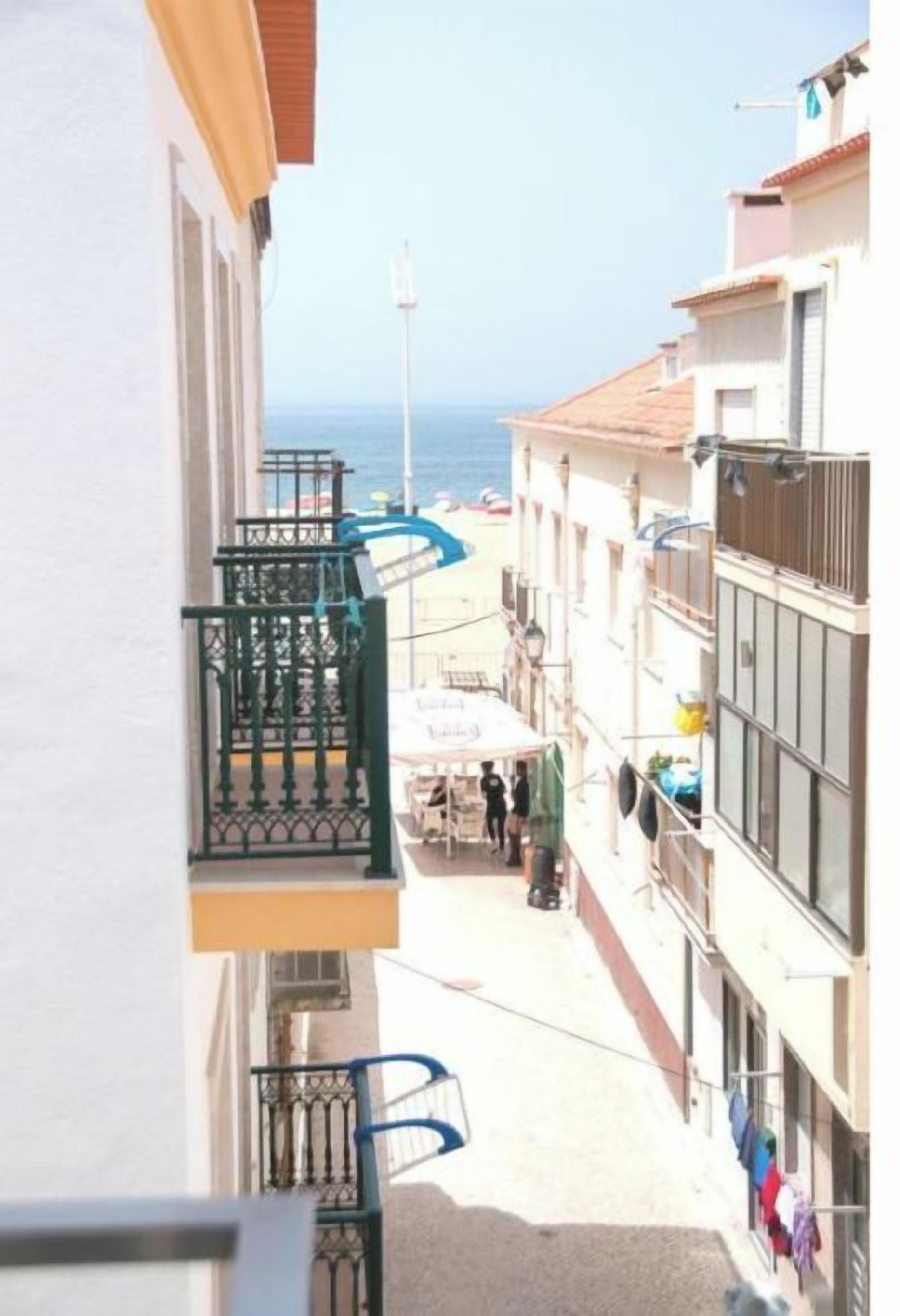 A narrow street with buildings on either side, a balcony with a metal railing, and a view of the sea in the background.