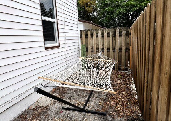 A hammock set up against a house with a wooden fence and a window.