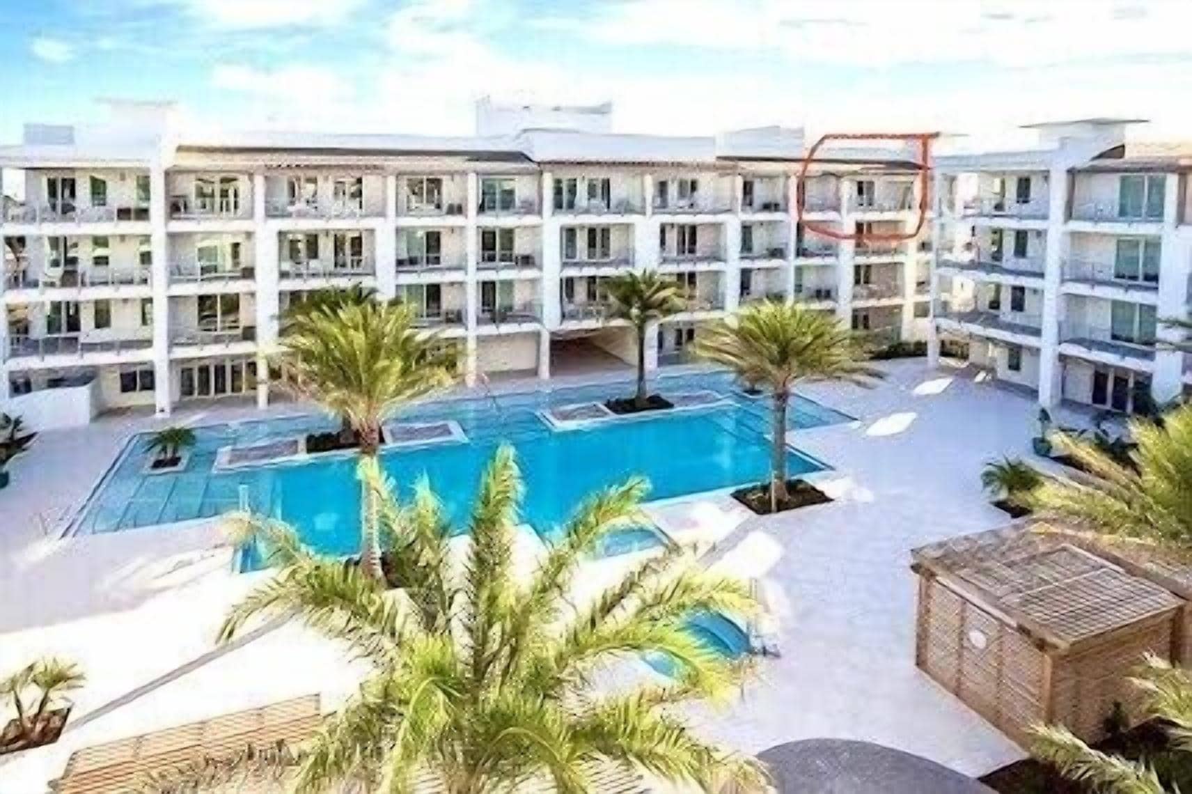 Ein mehrstöckiges Hotel mit einem zentral gelegenen Schwimmbecken, umgeben von Palmen und Liegestühlen.