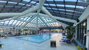 An indoor swimming pool with a glass ceiling, lounge chairs, and a caution sign for a wet floor.