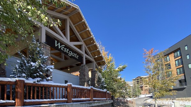 A building with a stone facade and a wooden deck, a clear blue sky, and a body of water with snow around it.
