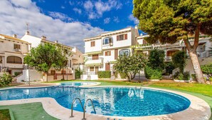 Une piscine à bord incurvé, entourée d’une terrasse verdoyante. On y voit des bâtiments blancs dotés de balcons, et un arbre au premier plan.