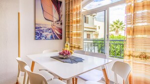 Un coin repas avec une table blanche, quatre chaises, un saladier de fruits et une bouteille de vin. Une grande fenêtre, ornée de rideaux, offre une vue sur un balcon et des immeubles.
