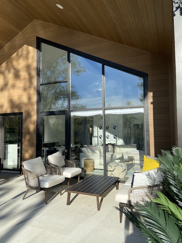 A modern outdoor seating area with wicker chairs and a glass table, large windows, and a wooden ceiling.