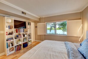 A bedroom with a large bed, a bookshelf, a window with blinds, and a view of a body of water.