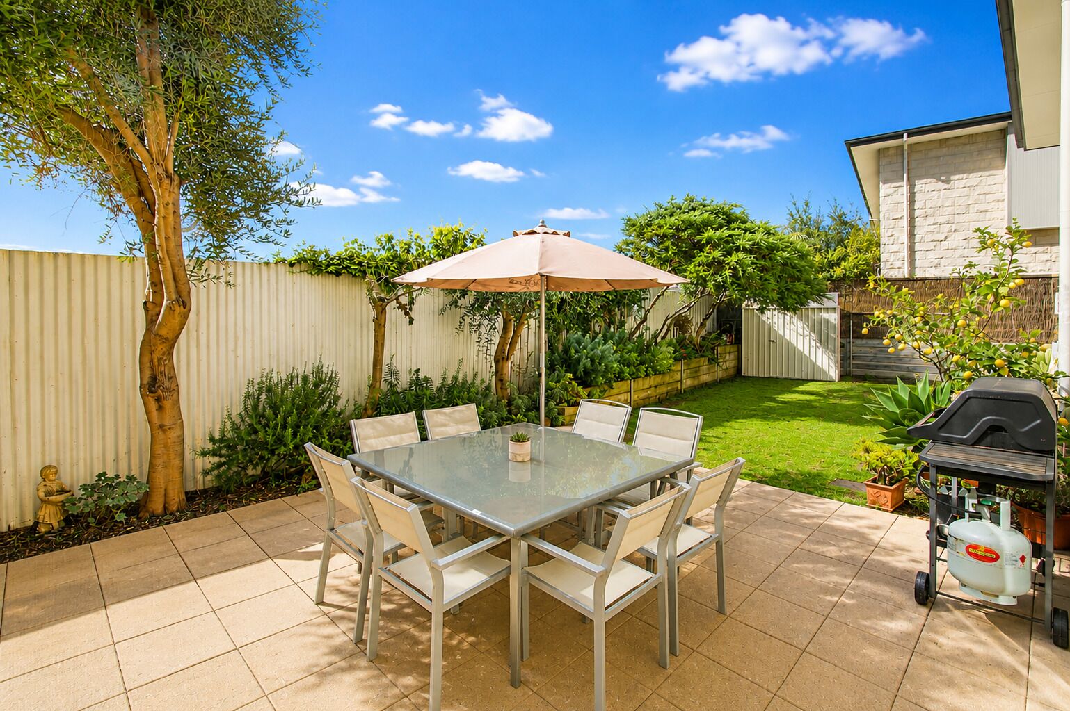 A patio with a glass table and chairs, a barbecue grill, and a fenced yard with various plants.