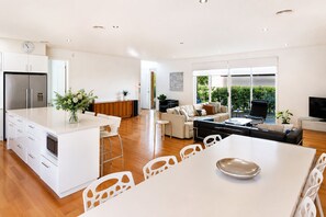 Een moderne keuken met een centraal eiland, een eethoek met een witte tafel en stoelen, een woonkamer met een bank en televisie, en een groot raam met uitzicht op groen.