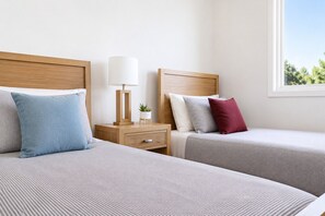 Quarto de hotel com duas camas, uma mesa de cabeceira com abajur e uma janela com vista para árvores.
