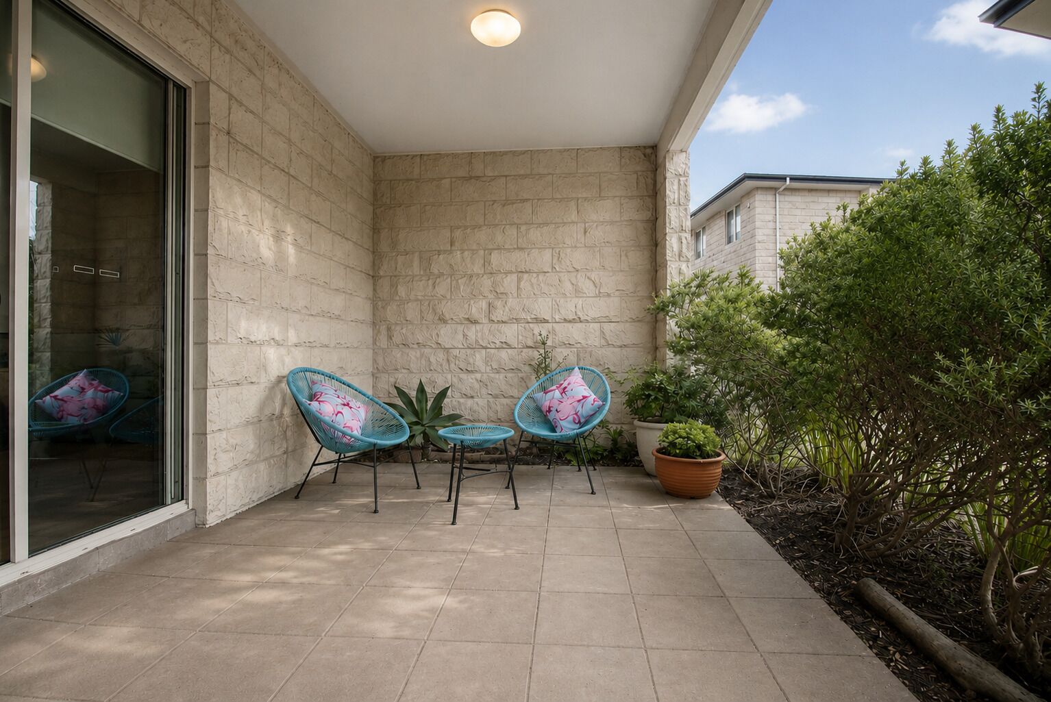 Ein überdachter Patio-Bereich mit Steinmauern, zwei blaue Stühle mit Blumenkissen, ein Blumentopf und Blick auf ein benachbartes Gebäude.