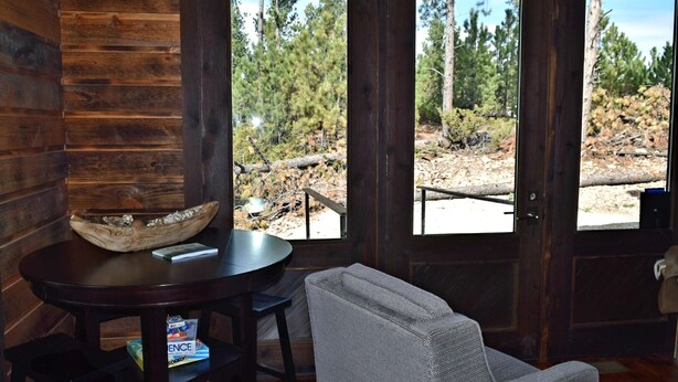 Un interior de madera con una mesa redonda, una silla acolchada y un amplio ventanal con vistas a una zona boscosa.